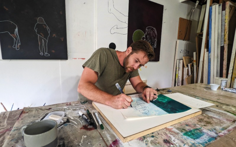 a male artist in a green shirt sketching a line drawing on a long wooden table. 