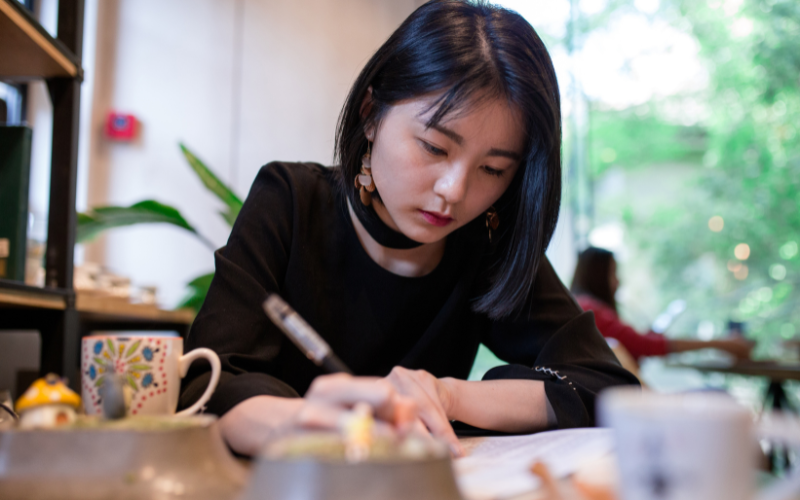 a light-skinned woman with short black hair writing on a table in a coffee shop