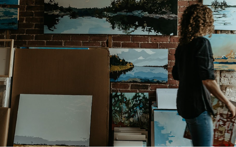 a woman in a black shirt and blue jeans is walking in front of a large brick wall covered with acrylic paintings of abstract representations of nature. While the paintings are focused and clear, she's in motion and is blurry