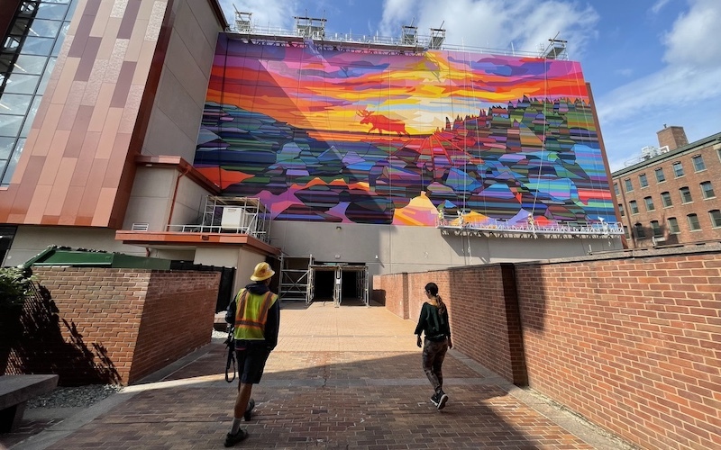 Construction worker and a woman walk up to a building with a colorful, geometric mural that is in process of being painted.