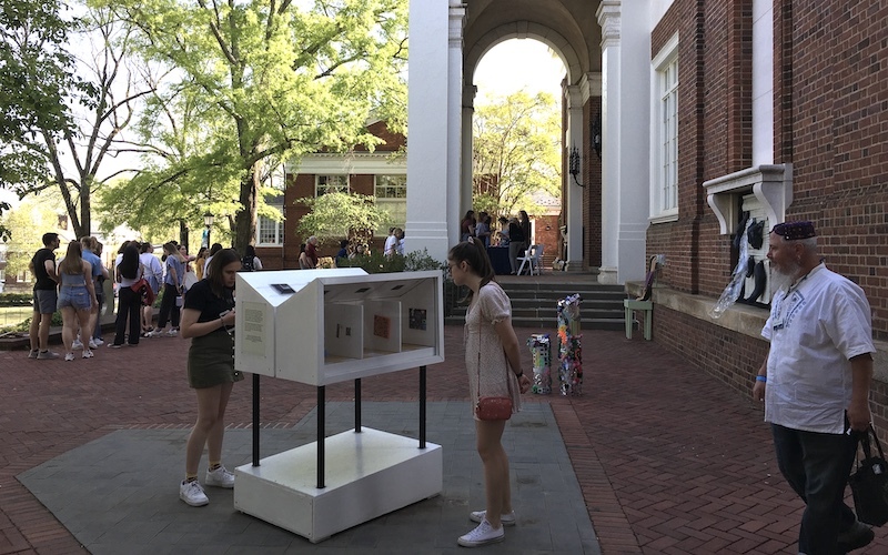 A group of people gather in a courtyard outside of a brick building. Two young women look at the miniature museum.