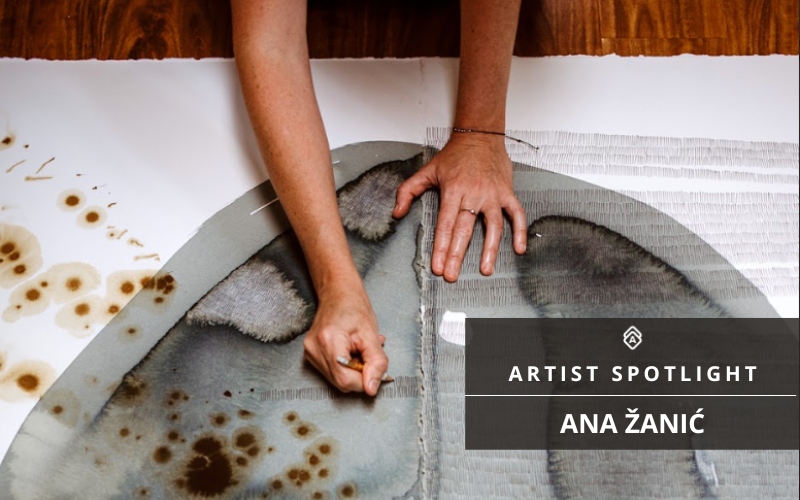 a promotional graphic for an artist spotlight, featuring an artist named Ana Žanić. The graphic shows the artist's hands working on a piece of paper or canvas with abstract watercolor. There's an overlay of text on the image that reads "ARTIST SPOTLIGHT ANA ŽANIĆ"