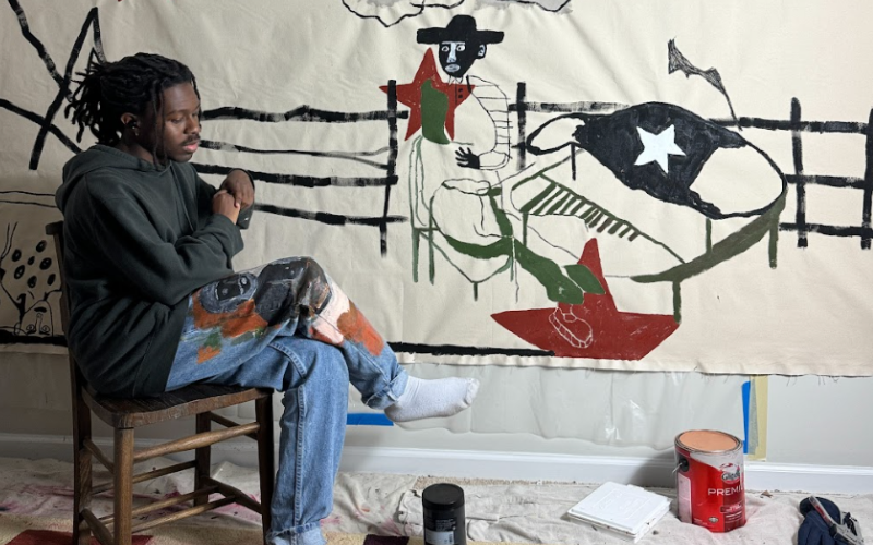 Artist known as Bhare sits with his legs crossed in a chair in front of a piece of his artwork. The artwork pictured is a large, canvas with an outline of an abstract piano, man with a cowboy hat, and red stars
