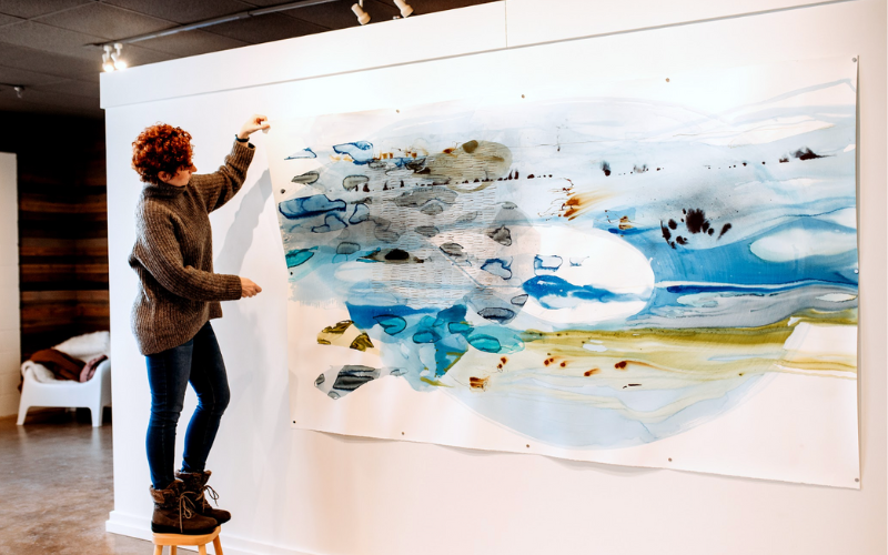 The image depicts artist Ana Zanic interacting with a large abstract painting. The painting is pinned to the wall and features a blend of blue, black, and brown hues with dynamic strokes and washes, suggesting a landscape or a water scene. The artist, who has curly red hair, is standing on a wooden stool and reaching out to touch or paint on the canvas. She is wearing a brown sweater and blue jeans