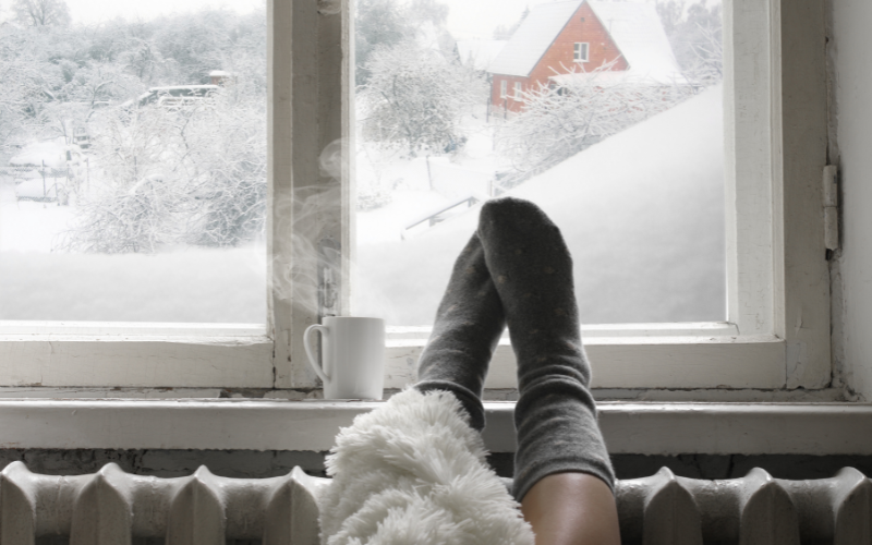 a light-skinned person's legs in socks and a cup of coffee by a frosty window. Outside you can tell it's just snowed