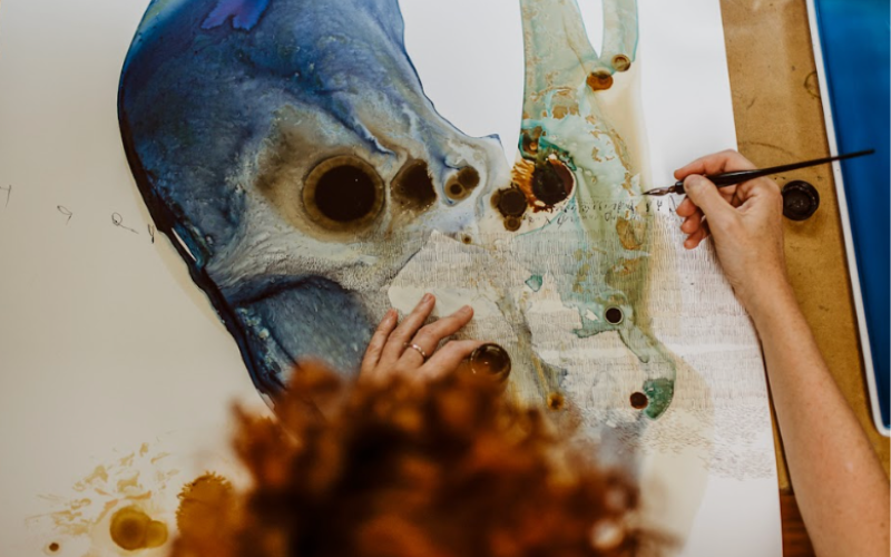 image shows a close-up view of artist Ana Zanic at work on a large abstract painting. The artist's hands are visible, one steadying the canvas and the other holding a paintbrush, applying detail to the artwork. The painting features organic shapes with a mix of blue and earth tones, and areas where the paint has created a blooming effect, often characteristic of watercolor techniques. The artist's curly red hair is partially visible in the foreground