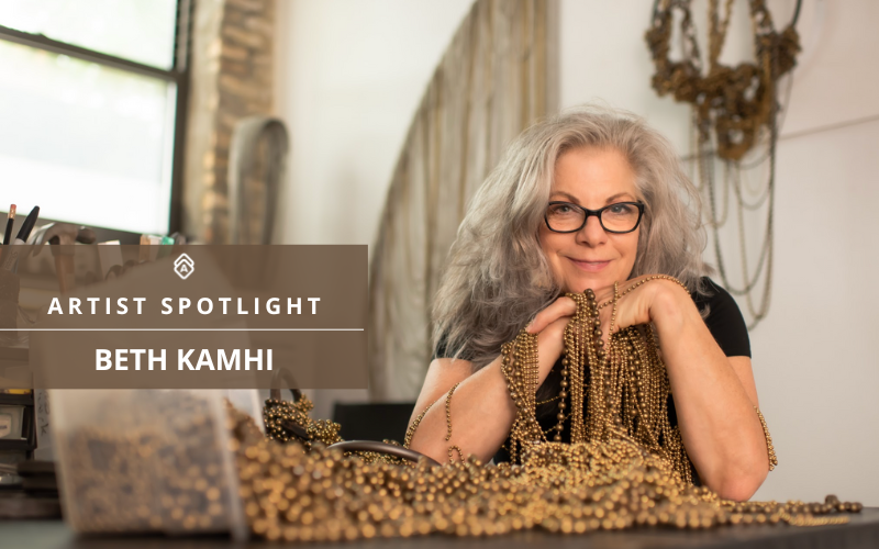 A light-skin toned woman with black rimmed glasses sitting at a table with her arms at her chin. steel ball chain beads are wrapped around her hands, arms, and spilling onto the table in front of her. Text on the image reads: Artist Spotlight Beth Kamhi