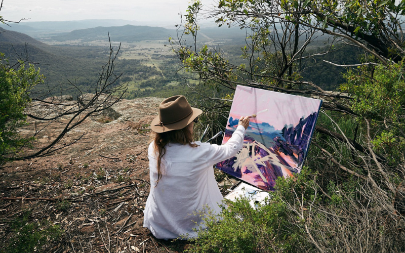 a woman with a hat painting en plein air overlooking a vast mountain landscape. 