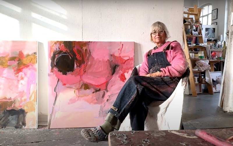 A woman sitting in front of two large pink abstract paintings on canvas. She's wearing a pink shirt, black overalls, and crocs and is sitting with one leg crossed over the other. 