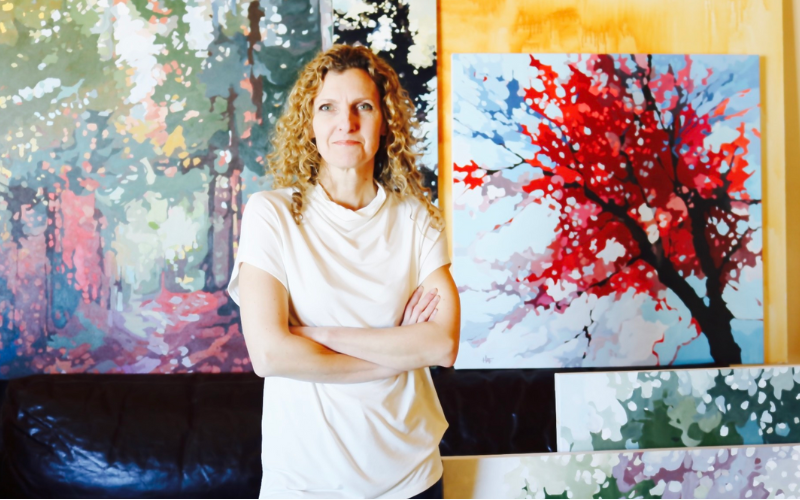 Holly Ann Friesen, with light-colored curly hair stands in front of a few large canvases of her abstract nature paintings with her arms crossed