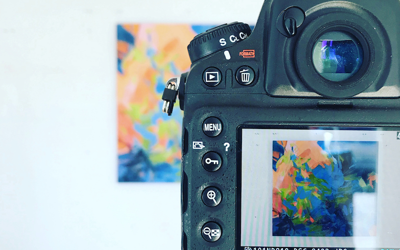 A close-up view of a digital camera capturing a photo of an artwork by Cameron Schmitz hanging in front of the cameras view. 