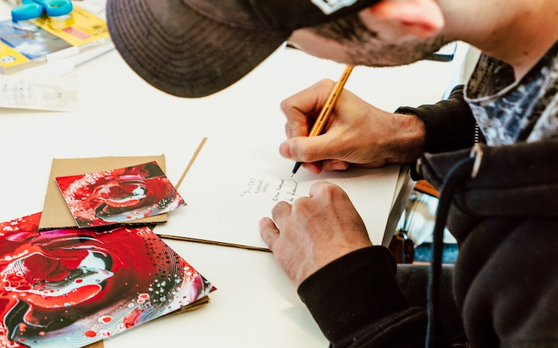 Artist signing art card: A male artist wearing a black hat leans over an art card, signing the back with a pencil.