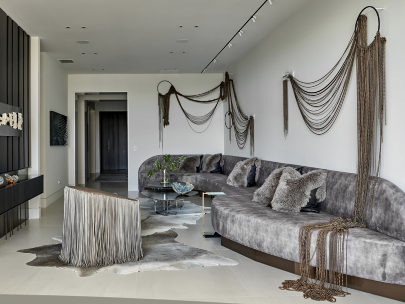 A large upscale living room with grey velvet couch, cow skin rugs, and Beth Kamhi's ball chain installation sculpture entitled Swag 3