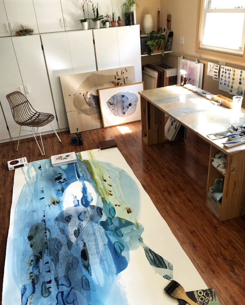 image provides a glimpse inside Ana Žanić's art studio. Natural light fills the room, highlighting a large abstract watercolor painting laid out on the floor, showcasing varying shades of blue and green with intricate line work. To the right, a wooden work table is seen, cluttered with art supplies, brushes, and palettes, suggesting an active and creative workspace. In the background, storage cabinets line the wall, with some artwork leaning against them, and a window offers a view outside, contributing to the room's bright ambiance